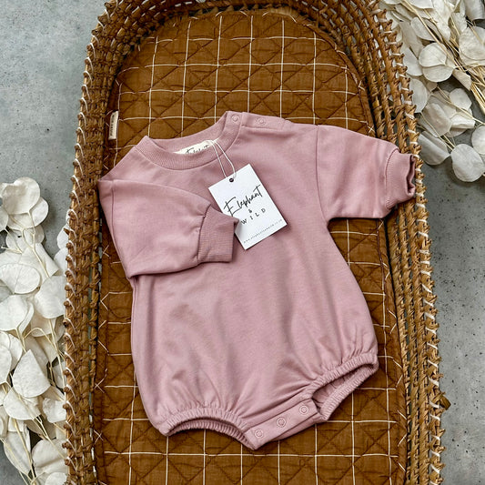 Pink baby romper with a tag on a woven mat with decorative flowers.