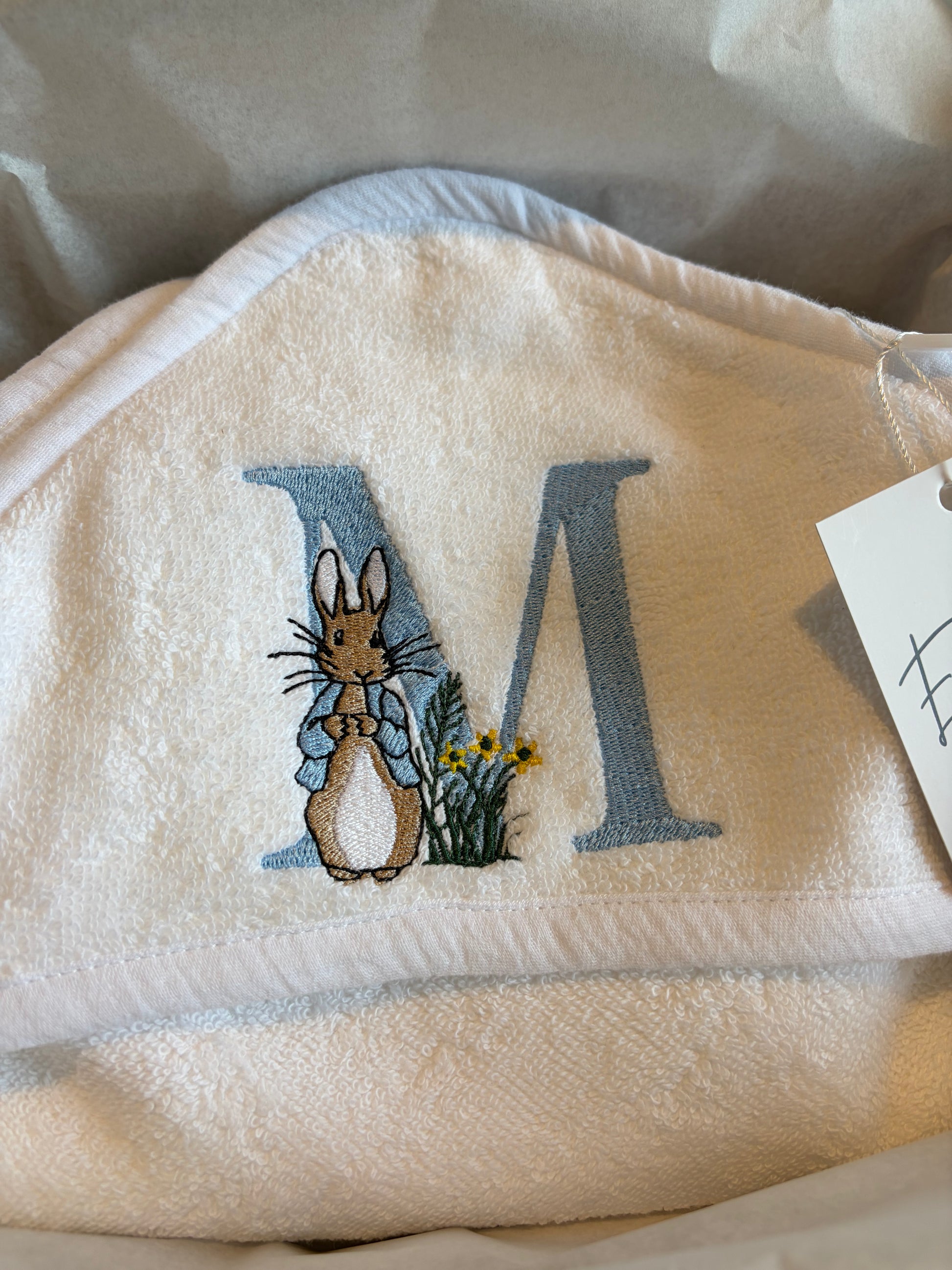 White personalised hooded baby towel with blue embroidered letter M featuring Peter Rabbit and yellow flowers.